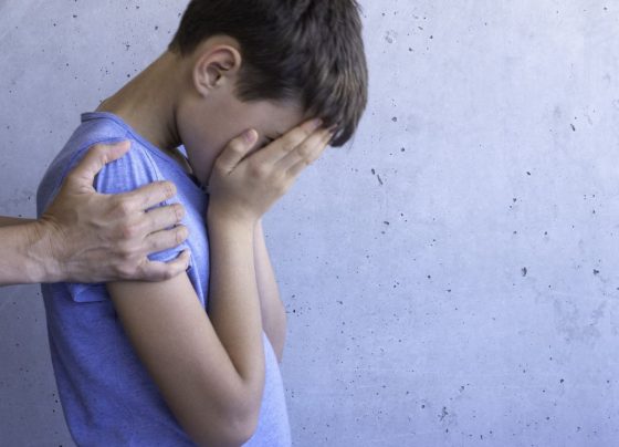 Foto colorida de criança cobrindo o rosto e sendo segurado por adulto. Imagem ilustrativa - Metrópoles