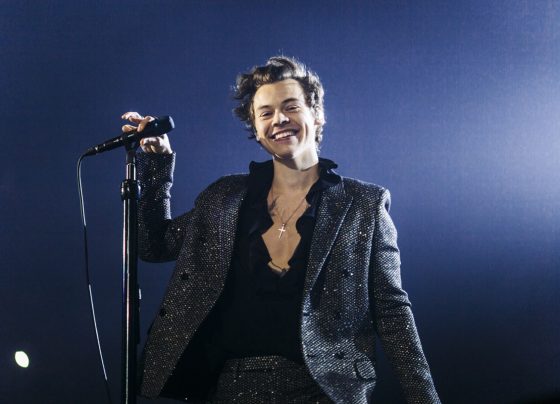 Imagem colorida de Harry Styles em show, sorrindo e usando roupa toda preta - Metrópoles