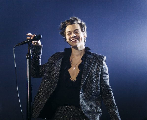 Imagem colorida de Harry Styles em show, sorrindo e usando roupa toda preta - Metrópoles